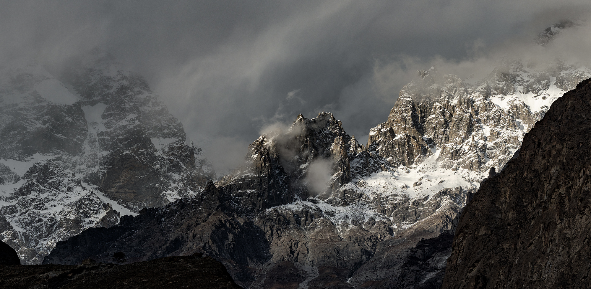 This photograph captures the tumultuous beauty of the Karakoram&rsquo;s peaks, where clouds and mountains engage in an eternal dance. Light breaks through the overcast sky, spotlighting the rugged cliffs and snow-dusted crevices, embodying the raw power of nature&rsquo;s artistry. The scene invites viewers to listen for the whispers of the wind that sculpts these giants—a symphony of natural forces at play.