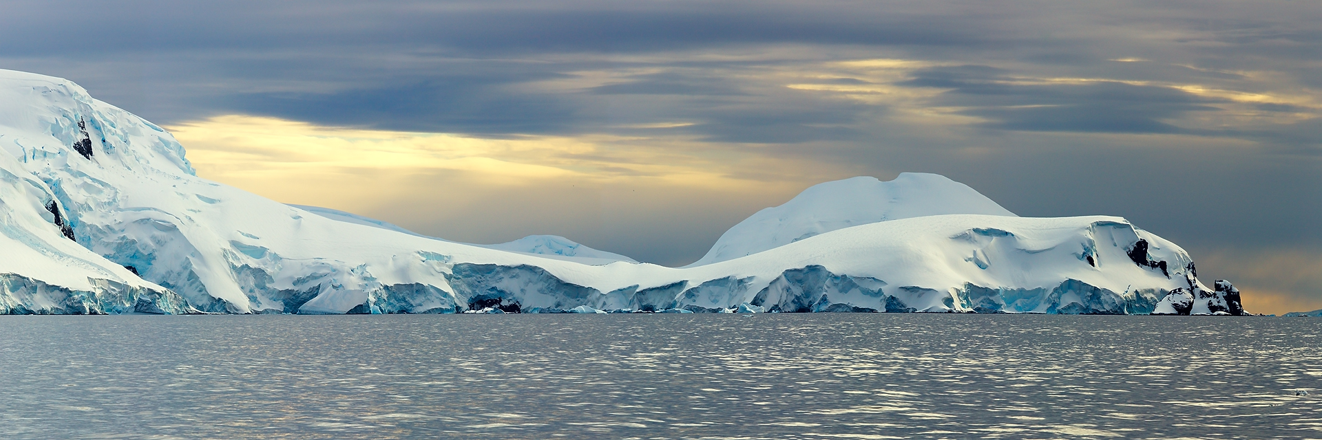 The tranquil waters of the polar sea reflect the soft glow of twilight, framing the majestic ice formations along the horizon. This panoramic image showcases the subtle interplay of color and light in the Antarctic wilderness, where the serene blues and whites of the ice contrast with the darkening sky. A testament to the quiet majesty of the Earth&rsquo;s southernmost landscapes, it is a serene backdrop for contemplation and a splendid addition to any art collection.