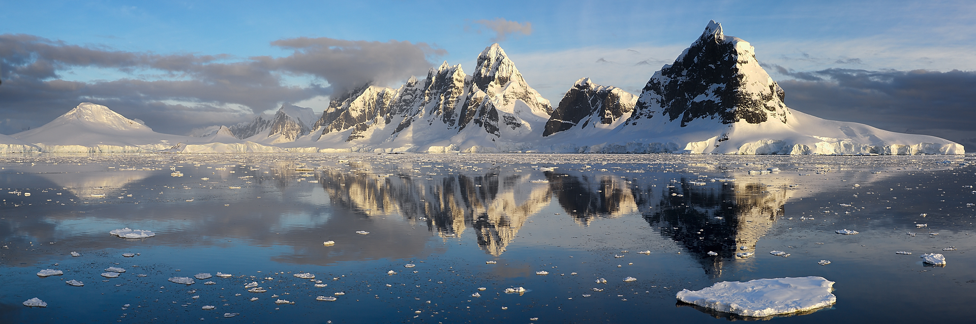 As the sun dips towards the horizon, its golden light bathes the Antarctic peaks, captured here in a moment of stunning clarity. The mirror-like waters offer a perfect reflection, doubling the majesty of the snow-capped mountains and the scattered ice that adorns the sea. This panoramic shot is a breathtaking showcase of the polar landscape, where the harmony of nature&rsquo;s grandeur and tranquility is eternally preserved.