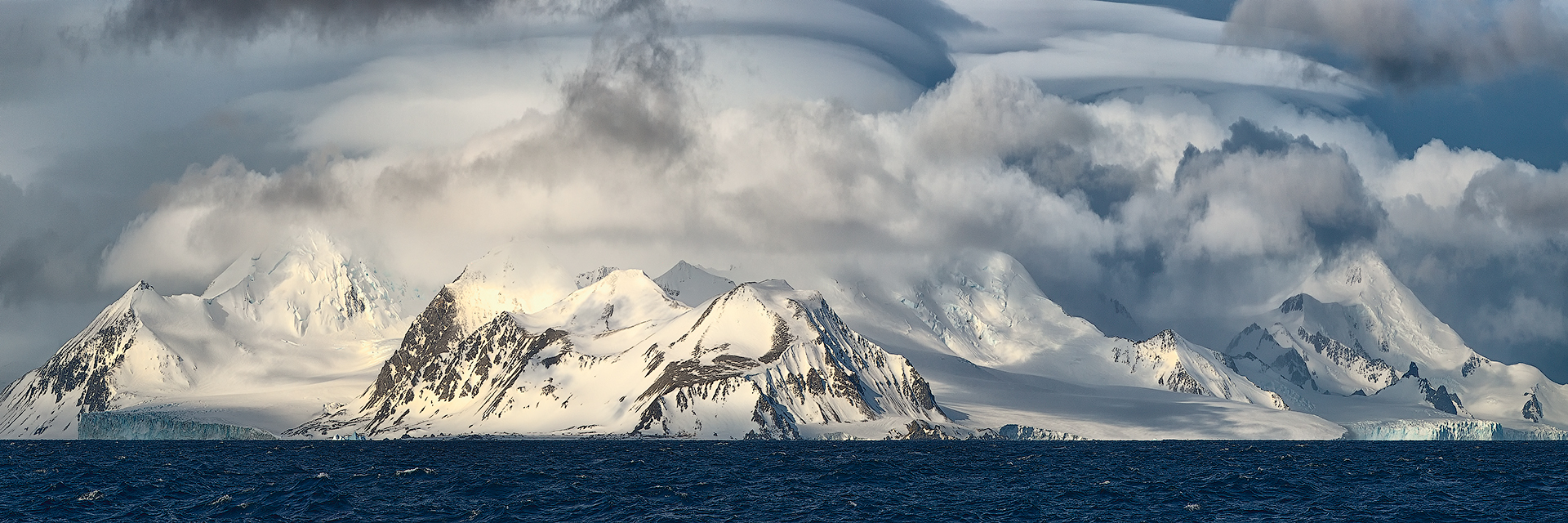 The dance of light and shadow plays across the Antarctic peaks, where the rugged mountains meet the tumultuous sky. The dark, rolling clouds, sculpted by the fierce polar winds, accentuate the majesty of the snow-covered summits. This panoramic image captures the contrast between the turbulent blue sea and the serene whiteness of the landscape, a testament to the raw and awe-inspiring power of Earth&rsquo;s southernmost wilderness.