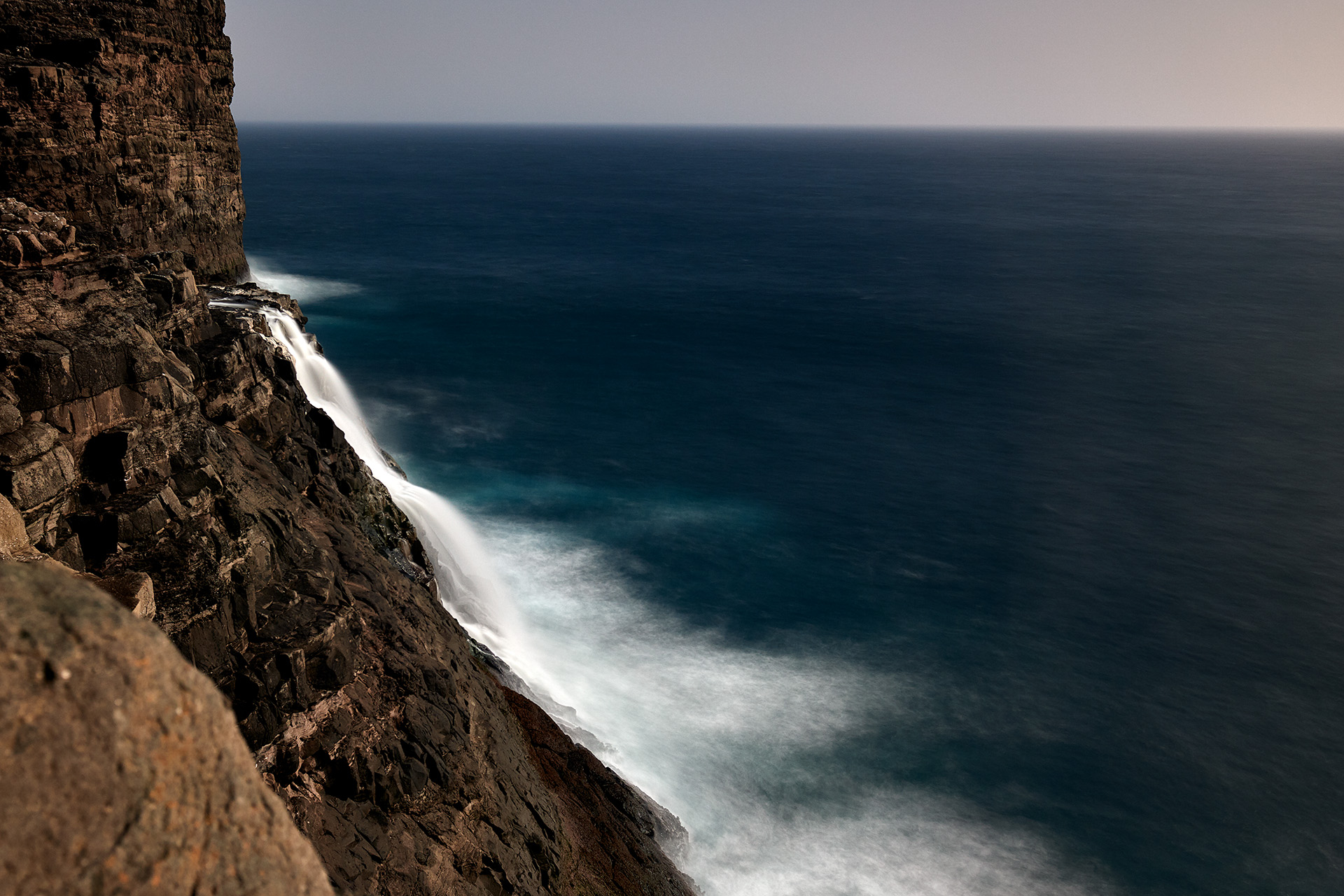 An extraordinary view of a waterfall plunging into the ocean from the steep cliffs of the Faroe Islands. The sheer force of the water creates a misty veil against the deep blue of the North Atlantic, illustrating the raw power and untouched beauty of the islands&rsquo; landscapes.